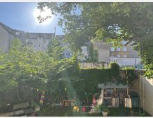 Hinterhof mit dichtem Grün, einer Wäscheleine mit bunten Wimpeln, einem Holzschuppen an einer mit Efeu bewachsenen Mauer und Spielzeug auf dem Boden. Im Hintergrund mehrstöckige Wohnhäuser unter einem sonnigen Himmel.