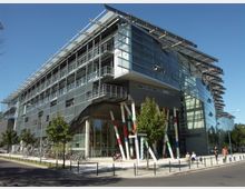 Modernes Büro- oder Universitätsgebäude mit einer Fassade aus Glas und Metall, markant durch schräg abstehende Sonnenblenden und bunte, schräg gestellte Säulen am Eingangsbereich. Im Vordergrund befinden sich Gehwege, Bäume und geparkte Fahrräder, während der Himmel klar und blau ist.