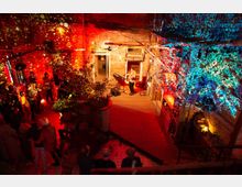 Ein Innenhof mit rotem Backstein und Holzplanken, beleuchtet von bunten Lichtprojektionen in Rot und Blau. Eine Person steht auf einer kleinen Bühne mit einem Keyboard, während ein Publikum umgeben von Pflanzen zuhört.