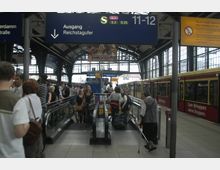 Bahnhofshalle mit großen Glasfenstern und ausgeschildertem Ausgang Richtung "Reichstagufer". Im Vordergrund eskalieren Menschen auf Rolltreppen, während rechts ein roter S-Bahn-Zug steht.