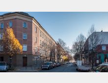 Straßenszene in einem Wohngebiet mit einem mehrstöckigen Gebäude aus rotem Putz auf der linken Seite, Bäumen entlang der Straße und parkenden Autos auf beiden Seiten. Der Himmel ist klar und die Bäume haben teilweise gelbes Herbstlaub.