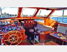 Innenraum einer Yacht mit Holzausstattung, einschließlich eines Steuerrads, einer Steuerkonsole mit Instrumenten sowie einer Sitzbank mit Tisch. Große Fenster bieten einen Ausblick auf das Wasser und die Landschaft im Hintergrund.