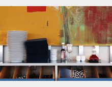 Ein Buffetbereich mit gestapelten weißen Tellern, dunklen Servietten, Gewürzen, einer Karaffe und Gläsern auf einer Ablage vor einem abstrakten, farbenfrohen Wandgemälde. Darunter sind Fächer mit Besteck und weiteren Utensilien zu sehen.