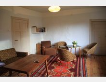 Wohnzimmer mit Holzboden und weiß gestrichenen Wänden, ausgestattet mit einem Holztisch, mehreren Sesseln und Stühlen mit gemusterten Bezügen. Ein bunter Teppich bedeckt den Boden, und ein kleiner runder Tisch mit einer Vase mit Blumen steht in der Mitte; ein Bücherregal hängt an der Wand.