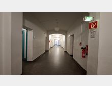 Langer, weiß gestrichener Innenflur mit gewölbter Decke und runden Deckenleuchten, entlang der Wände mehrere Türöffnungen und Durchgänge mit Bögen. An der rechten Wand befinden sich ein Feuerlöscher sowie ein grünes Notausgangsschild und ein rotes Brandschutzsymbol.