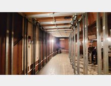 Ein schmaler Raum mit Holzverkleidung und mehreren vertikalen Metallstreben, die die Decke stützen. Der Boden besteht aus Holzdielen, und an der Decke befinden sich Schienen mit Rollen.