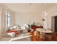 Helles Wohnzimmer mit Stuckdecke, einem großen Fenster mit weißen Vorhängen und Holzdielenboden. Im Raum stehen ein cremefarbenes Sofa mit orangener Decke, ein Glas-Couchtisch, eine halbrunde Stehlampe, sowie ein Esstisch aus Marmor mit Holzstühlen; im Hintergrund eine Kommode mit Dekoobjekten und Pflanzen.