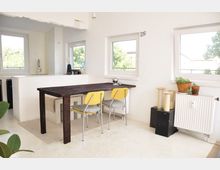 Minimalistisch eingerichteter Essbereich in einer Wohnung mit weiß gestrichenen Wänden, großen Fenstern, einem dunklen Holztisch und zwei gelben Stühlen. Im Hintergrund ist eine offene Küche mit Spülbecken sichtbar, und auf der rechten Seite befinden sich Pflanzen und Dekorationen auf einem Heizkörper.