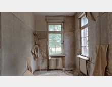 Ein leerer, verfallener Raum mit abblätternder Tapete und freiliegender Backsteinmauer. In der Mitte befindet sich ein Fenster mit blickbarer Verglasung, darunter ein alter Heizkörper; der Boden ist mit Schmutz und losen Materialien bedeckt.