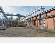 Ehemalige Industrieanlage mit Backsteingebäuden und großen Fenstern, ergänzt durch eine Stahlstruktur mit Laufkran. Der Außenbereich ist weitläufig mit Schotterboden und diversen abgelagerten Baumaterialien, während im Hintergrund Bäume sichtbar sind.
