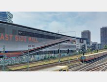 Außenansicht der East Side Mall in Berlin mit grauer Fassade und großem, orangefarbenem Schriftzug, umliegenden Hochhäusern und einem angrenzenden Bahngleis mit einem vorbeifahrenden Zug im Vordergrund. Die Umgebung ist urban, mit Graffiti an den Wänden entlang der Gleise.