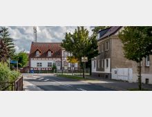 Straßenszene in einem Wohngebiet mit einem zweistöckigen Fachwerkhaus mit rotem Ziegeldach im Hintergrund, umgeben von Bäumen und Schildern, die Verkehrshinweise und Ortsangaben geben. Rechts im Bild stehen weitere Wohnhäuser mit schlichten Fassaden.