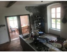 Große, rustikale Küche mit freistehender Kücheninsel, umgeben von Geschirr und Utensilien. Im Hintergrund ein Essbereich mit Holztisch und Stühlen, abgetrennt durch eine Türöffnung; große Fenster lassen Tageslicht in den Raum.