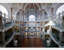 Großzügiges, historisches Treppenhaus mit symmetrischem Design, verziert mit geschwungenen Geländern und Säulen. Gewölbedecke, Rundbogenfenster und gefliester Boden prägen die Architektur des Innenraums.