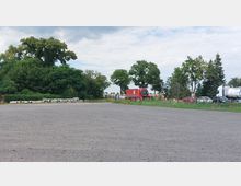 Großer, leerer Schotterplatz mit vereinzelten Betonrohren am Rand, umgeben von Bäumen und Sträuchern. Im Hintergrund parken mehrere Lastwagen und Autos neben einer kleinen Wiese und einem Verkehrszeichen.