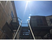 Metalltreppe zwischen zwei Gebäuden mit Putz- und Backsteinfassaden unter klarem Himmel; Sonnenstrahlen scheinen von oben herab.