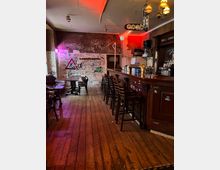 Innenraum einer rustikalen Bar mit Holzdielenboden, langen Tresen mit Barhockern und Regalen mit Flaschen. Die Wände sind dicht mit Graffiti und Stickern bedeckt, dazu hängen Schilder und warme Deckenlampen, teils in farbigem Licht.