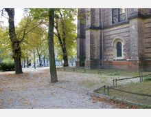 Außenansicht eines historischen Backsteingebäudes, vermutlich einer Kirche, mit großen Spitzbogenfenstern und dekorativen Ziegelmustern. Im Vordergrund befindet sich ein Baum bestandener Platz mit herbstlichem Laub, im Hintergrund sind parkähnliche Wege und umliegende Wohnhäuser zu erkennen.