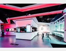 Modernes Café oder Restaurant mit futuristischem Design und offener Theke. Die Innenraumgestaltung umfasst weiße geometrische Elemente, rote und pinke LED-Beleuchtung sowie eine Sitzbereich mit schwarzen Tischen und Stühlen, ergänzt durch große Fensterfronten, die Tageslicht einlassen.