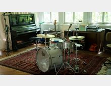 Ein Raum mit Musikinstrumenten, darunter ein Schlagzeug auf einem roten Teppich und ein Klavier im Hintergrund. Im Raum befinden sich zudem Gitarren, ein Tisch mit Musikgeräten und Kabeln sowie große Fenster, die Tageslicht hereinlassen.