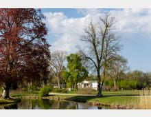 Weitläufiger Park mit einem kleinen See im Vordergrund, umgeben von Bäumen in verschiedenen Farben, darunter ein Baum mit roten Blättern. Im Hintergrund steht ein weißes Gebäude im klassizistischen Stil mit Säulen, eingebettet in eine gepflegte Grünfläche.