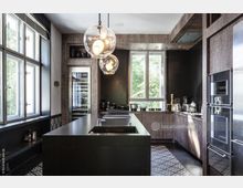 Moderne Küche mit dunklen Holzschränken, einer großen zentralen Kücheninsel mit integriertem Spülbecken und drei kugelförmigen Hängeleuchten. Große Fenster lassen Tageslicht herein, während Edelstahlgeräte und dekorative Fliesen den Raum ergänzen.