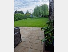 Blick von einer gepflasterten Terrasse in einen großen, gepflegten Garten mit Rasenfläche, hoher Hecke am Rand und einer einzelnen Schaukel. Im Hintergrund steht ein großer, herabhängender Baum, rechts ist die Außenwand eines Holzgebäudes mit Pflanzkübel zu sehen.