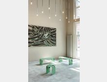 Minimalistisch eingerichteter Innenraum mit beigefarbenen Wänden und einer großen abstrakten Kunstinstallation an der Wand. Im Vordergrund stehen drei mintgrüne, geometrische Sitzmöbel aus perforiertem Material; von der Decke hängen schlanke, vertikale Lampen, und rechts sind hohe Fenster und ein Heizkörper zu sehen.