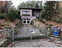 Wohnhaus in ländlicher Umgebung mit einer Auffahrt und Steingarten. Die Einfahrt ist durch ein Metalltor und eine mit Natursteinen gemauerte Begrenzung eingefasst, umgeben von Bäumen und herbstlichem Laub.