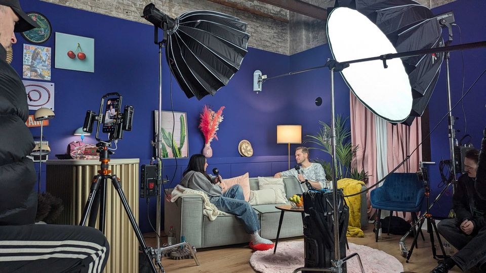 Studio-Set in einer hohen Industriehalle mit blau gestrichenen Wänden, grauem Sofa, Stehlampe, Vorhängen und dekorativen Bildern. Im Vordergrund stehen Kamera auf Stativ sowie große Softbox-Leuchten und weiteres Filmequipment auf Holzboden mit rundem Teppich und kleinem Tisch.