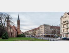Eine neugotische Backsteinkirche mit zwei spitzen Türmen steht links auf einem parkähnlichen Rasenbereich. Rechts im Bild befinden sich mehrstöckige Wohn- und Geschäftshäuser entlang einer Straße mit parkenden Autos und Geschäften im Erdgeschoss.