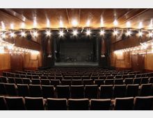 Innenraum eines Theaters mit leeren Sitzreihen, einer großen Bühne im Hintergrund und holzgetäfelten Wänden. Die Decke und Wände sind mit warmem Licht beleuchtet, das die Umgebung hell erstrahlen lässt.