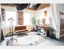 Ein heller Raum mit unverputzten Ziegelwänden, großen Fenstern und einem Frisiertisch mit Spiegel. Der Raum ist mit einem Friseurstuhl, einer Sitzbank, einer Kleiderstange, Topfpflanzen und einem orientalischen Teppich eingerichtet.