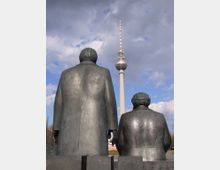 Bronzeskulpturen von zwei sitzenden Figuren, die mit dem Rücken zur Kamera gewandt sind, im Vordergrund; im Hintergrund der Berliner Fernsehturm unter einem bewölkten Himmel.