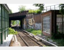 Bahnstrecke mit zwei Gleisen, die unter einer rostigen Eisenbahnbrücke hindurchführen, umgeben von Backsteingebäuden mit Graffiti und großen Fenstern. Im Vordergrund sind grüne Pflanzen und ein überdachter Gehweg auf der linken Seite sichtbar.