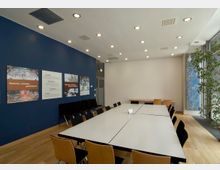 Moderner Konferenzraum mit rechteckigen Tischen in der Mitte und Holzstühlen mit schwarzen Sitzflächen. Links eine dunkelblaue Wand mit mehreren Postern, rechts große Fenster mit Pflanzen im Vordergrund, hellem Holzfußboden und weißer Decke mit eingebauten Leuchten.