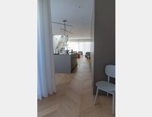 Modernes Wohnzimmer mit offener Küche und minimalistischer Inneneinrichtung. Zu sehen sind helle Holzböden, eine Kücheninsel, eine freitragende Treppe mit Metallgeländer und bodentiefe Fenster mit transparenten Vorhängen.