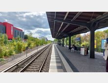 Überdachter Bahnsteig eines Bahnhofs mit Sitzbänken und Personen, die auf Züge warten. Links sind Bahngleise, Vegetation und ein modernes, rotes Gebäude sichtbar, im Hintergrund ziehen Wolken über den Himmel.