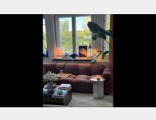 Wohnzimmer mit einem großen, rostfarbenen Sofa, einem runden Beistelltisch mit Kerze und einem Couchtisch mit Büchern und Blumen. Im Hintergrund sind große Fenster mit Blick auf Bäume und eine Stadtlandschaft, dekoriert mit Tischlampen, Kunstobjekten und Pflanzen.