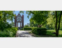 Blick auf ein historisches neugotisches Backsteintor mit turmartigen Pfeilern und glasierten Verzierungen, umgeben von einer grünen Parklandschaft mit dichtem Laub und einem gepflasterten Weg im Vordergrund.