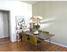 Esszimmerbereich mit einem rechteckigen Tisch aus Holz und vier grünen Stühlen. Über dem Tisch hängt eine blumenförmige Pendelleuchte, auf dem Tisch stehen eine Vase mit Lilien und Kerzen. Im Hintergrund ein Bücherregal mit Kunstwerken und eine geöffnete Tür, Holzdielenboden sichtbar.