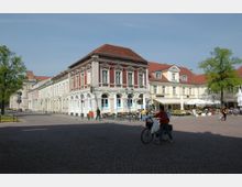 Historischer Platz mit breitem, gepflastertem Boden, umgeben von restaurierten Gebäuden im klassischen Stil mit roten Ziegeldächern und verzierten Fassaden. Im Vordergrund sind Personen und ein Cafébereich mit Sonnenschirmen sichtbar, während links und rechts Bäume stehen.