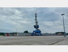 Ein industrielles Hafengelände mit einem großen blauen Hafenkran auf einer betonierten Fläche. Im Hintergrund sind Betonwände, Bäume und Lagerhügel zu sehen, unter bewölktem Himmel.