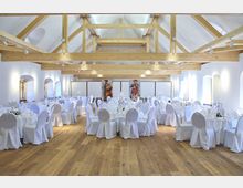 Ein heller Festsaal mit Holzboden, sichtbaren Holzträgern unter einer hohen Decke und weiß eingedeckten Tischen sowie Stühlen mit Hussen. Große Fenster in weißer Wandgestaltung lassen viel Tageslicht herein, und an der Rückwand befinden sich dekorative Figuren.