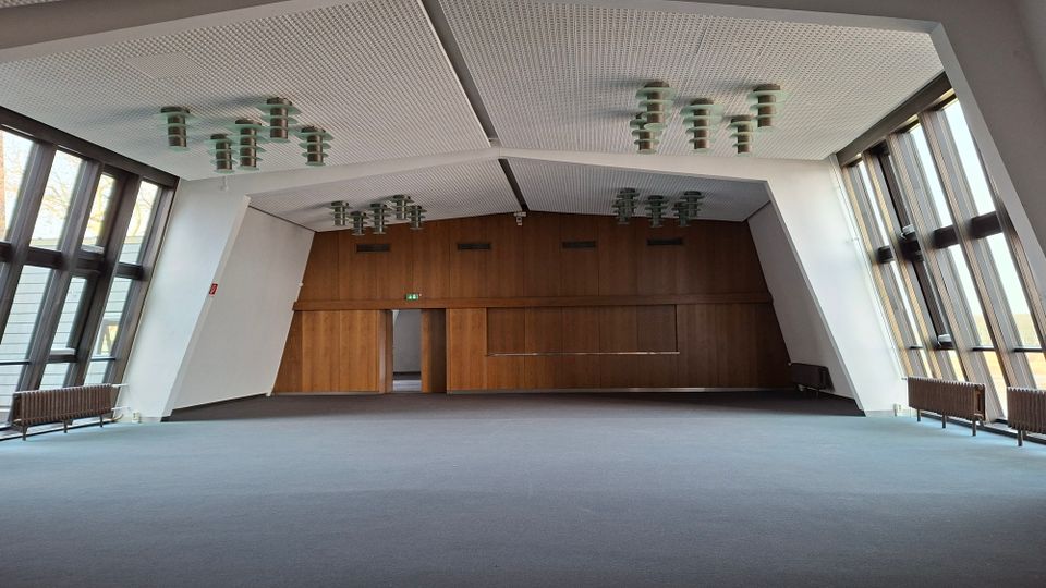 Großer, leerer Saal mit hoher, geometrisch gegliederter Decke und mehreren runden Deckenleuchten. An beiden Seiten verlaufen raumhohe Fensterfronten; die Stirnwand ist mit Holzvertäfelung und Türen gestaltet.