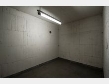 Kleiner leerer Raum mit unbehandelten Betonwänden, grauem Betonboden und einer rechteckigen Deckenlampe mit kaltem Licht. Eine runde Öffnung in der Wand sowie eine schlichte, funktionale Gestaltung sind sichtbar.