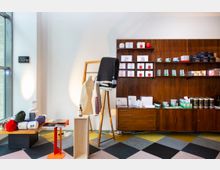 Innenansicht eines modernen Shops mit dekorativer Holzregalwand, auf der verschiedene Produkte wie Dosen, Boxen und Behälter angeordnet sind. Im Vordergrund stehen ein Kleiderständer mit Textilien, ein kleines Holzmöbel und ein Tisch mit bunten Accessoires, auf einem Teppich mit geometrischem Muster.