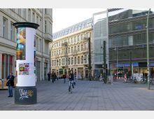 Straßenszene in einer urbanen Umgebung mit einem breiten, gepflasterten Fußgängerbereich. Links stehen historische Gebäude mit Ornamentfassaden, rechts befindet sich ein moderner Glasbau. Im Vordergrund ist eine Litfaßsäule mit Plakaten, und mehrere Menschen gehen oder fahren Fahrrad.