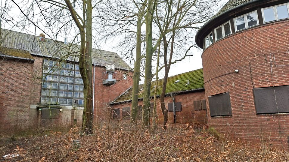Verlassen wirkender Backstein-Gebäudekomplex mit einem runden Anbau rechts und einem großen Glasbausteinfenster links, mehrere Fenster und Türen sind mit Platten verschlossen. Davor stehen kahle Bäume und dichter Bewuchs auf einem ungepflegten Gelände.