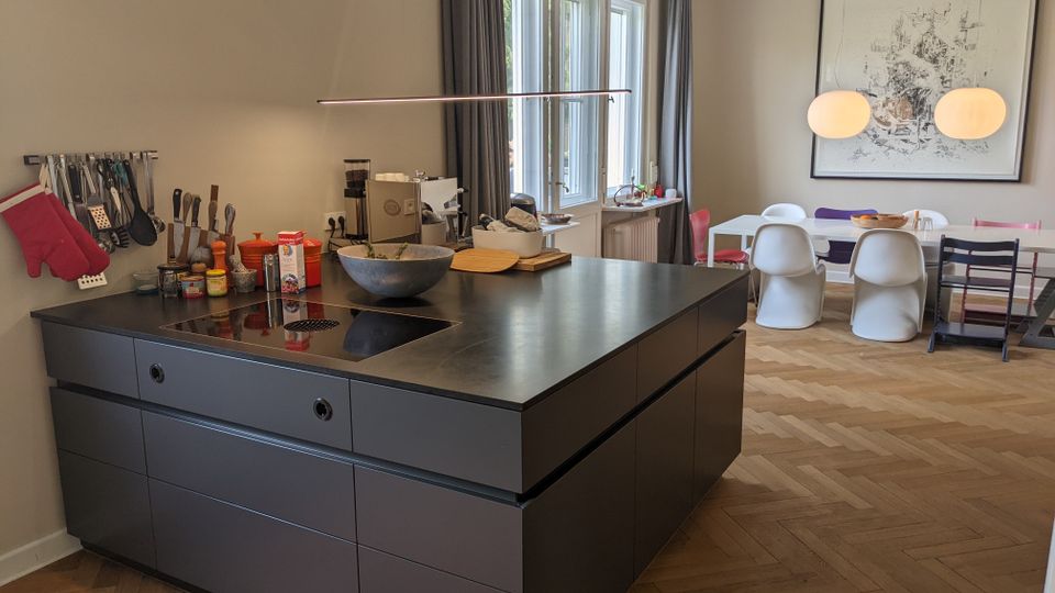 Offene Wohnküche mit großer dunkler Kücheninsel und integrierter Kochfläche, daneben eine Wand mit aufgehängten Küchenutensilien. Im Hintergrund ein Essbereich mit langem Tisch, gemischten Stühlen, zwei runden Pendelleuchten, großem Wandbild sowie hohen Fenstern mit Vorhängen über Fischgrätparkett.